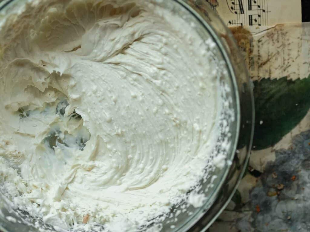 A glass bowl filled with whipped vegan cream sits on a floral and music sheet background. The cream is smooth, with some uneven textures and swirls, showing signs of being recently mixed or whipped.